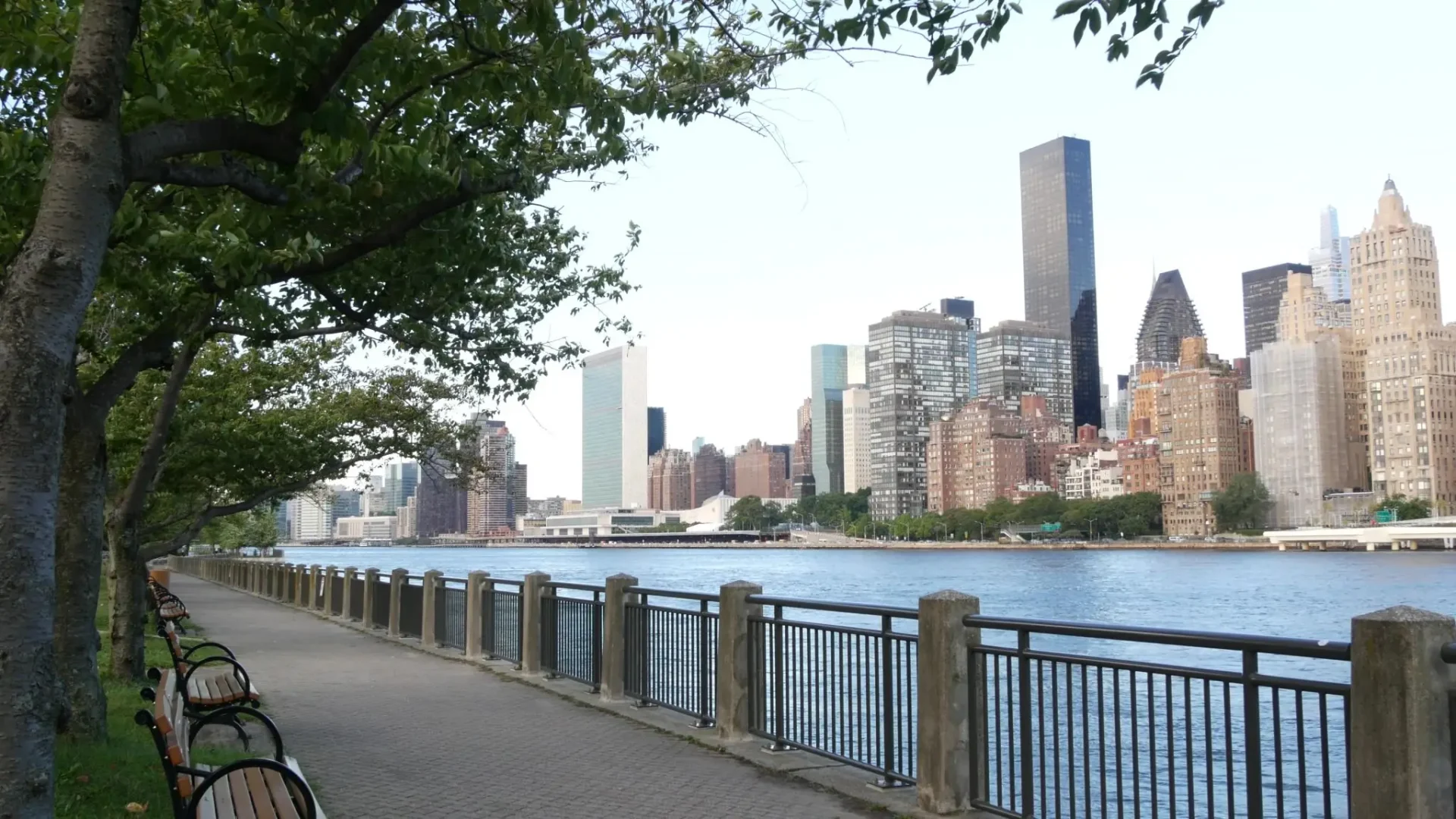 City skyline viewed from a riverside walkway with trees and railing.